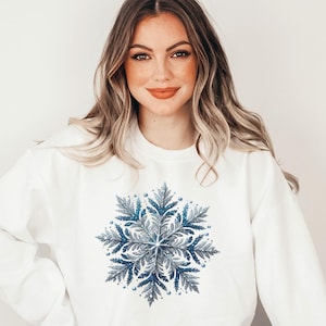 Op de afbeelding: Witte sweatshirt met een gedetailleerd sneeuwvlokontwerp in blauw en grijs. De sneeuwvlok heeft een symmetrisch, botanisch geïnspireerd patroon met kleine, ronde accenten. De sweatshirt is een casual kledingstuk met lange mouwen.