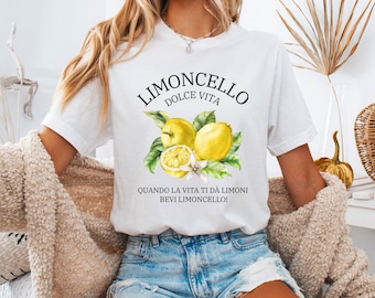 May include: A white short-sleeved t-shirt featuring a graphic of yellow lemons with green leaves. The text reads LIMONCELLO DOLCE VITA and QUANDO LA VITA TI DÀ LIMONI BEVI LIMONCELLO! The shirt is paired with light blue denim shorts.