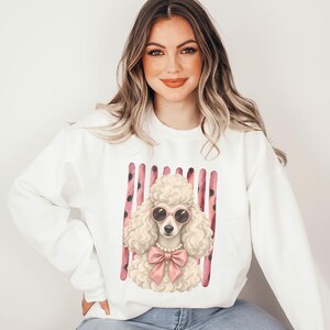 Könnte beinhalten: Ein weißes Langarm-Sweatshirt mit Rundhalsausschnitt und einem zentralen Aufdruck eines cremefarbenen Pudels, der eine rosa Sonnenbrille, eine Perlenkette und eine große rosa Schleife trägt. Der Hintergrund des Motivs besteht aus rosa Streifen.