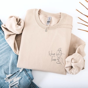 Könnte beinhalten: Ein beigefarbenes Sweatshirt mit Rundhalsausschnitt und einer schwarzen Strichgrafik auf der unteren rechten Seite. Das Motiv zeigt eine Hand, die ein Weinglas mit einem Herz darüber hält, sowie den Schriftzug Wine Time in Schreibschrift.