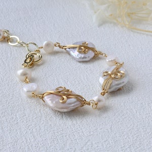 May include: A delicate pearl bracelet with gold wire accents. The bracelet features three large, irregularly shaped pearls wrapped in gold wire with intricate designs. The pearls are a soft white colour and the gold wire is a bright, shiny gold.