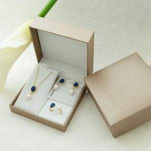K&ouml;nnte beinhalten: Ein Schmuckset in einer beigefarbenen Schachtel. Der Schmuck umfasst eine Halskette mit einem blauen Steinanh&auml;nger, ein Paar Ohrringe mit blauen Steinanh&auml;ngern und einen Ring mit einem blauen Stein. Der Schmuck wird auf einem wei&szlig;en Hintergrund pr&auml;sentiert.