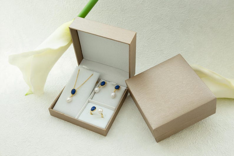 K&ouml;nnte beinhalten: Ein Schmuckset in einer beigefarbenen Schachtel. Der Schmuck umfasst eine Halskette mit einem blauen Steinanh&auml;nger, ein Paar Ohrringe mit blauen Steinanh&auml;ngern und einen Ring mit einem blauen Stein. Der Schmuck wird auf einem wei&szlig;en Hintergrund pr&auml;sentiert.