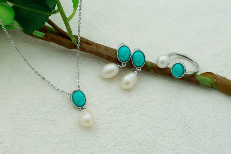 K&ouml;nnte beinhalten: Ein Schmuckset aus T&uuml;rkis und Perlen mit einer Halskette, Ohrringen und einem Ring. Die T&uuml;rkis-Steine sind oval geformt und in Silber gefasst.