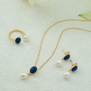K&ouml;nnte beinhalten: Eine goldene Halskette mit einem blauen ovalen Anh&auml;nger und einem Perlen-Tropfen, dazu passende Ohrringe mit blauen ovalen Steinen und Perlen-Tropfen sowie ein goldener Ring mit einem blauen ovalen Stein und einer Perle.
