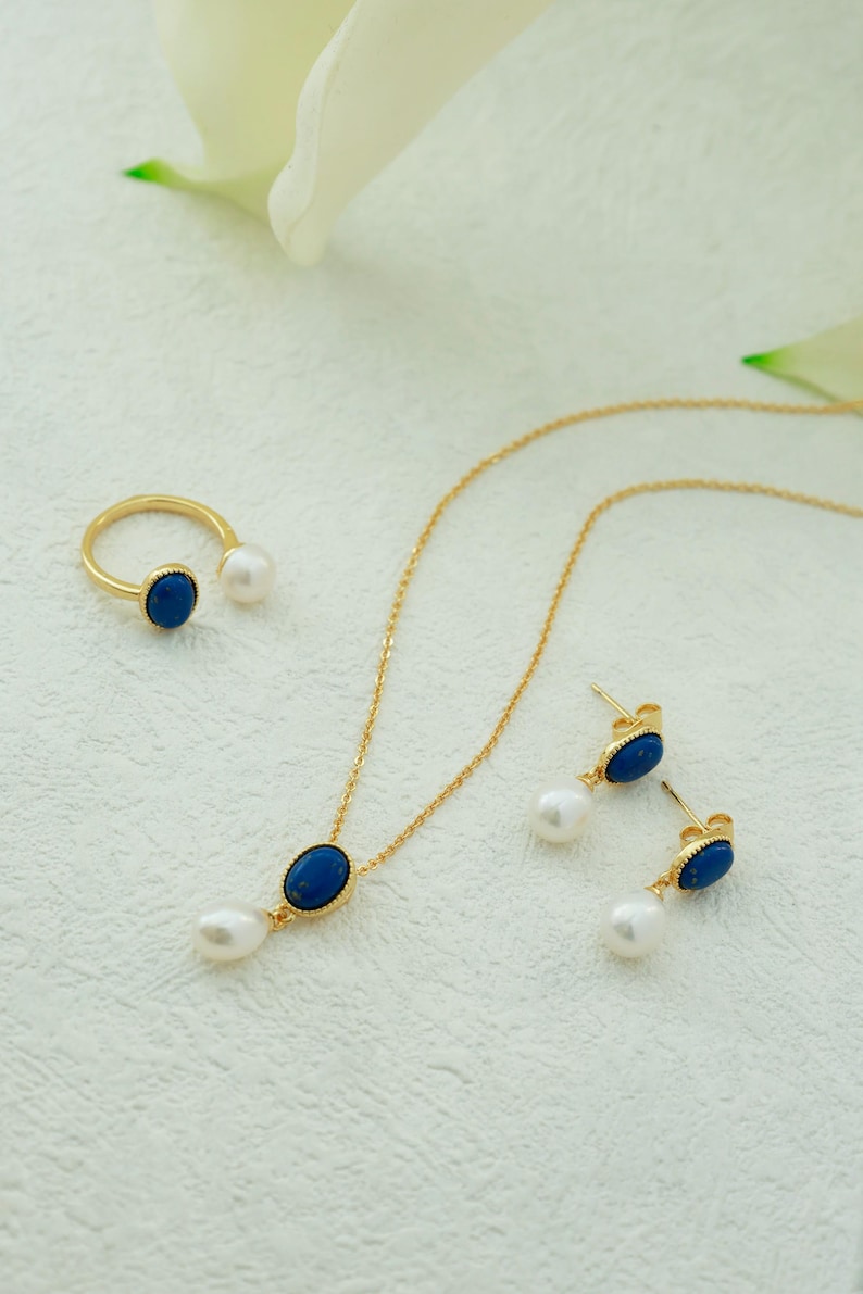 K&ouml;nnte beinhalten: Eine goldene Halskette mit einem blauen ovalen Anh&auml;nger und einem Perlen-Tropfen, dazu passende Ohrringe mit blauen ovalen Steinen und Perlen-Tropfen sowie ein goldener Ring mit einem blauen ovalen Stein und einer Perle.