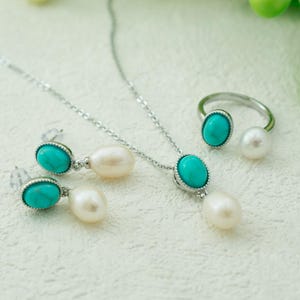 K&ouml;nnte beinhalten: Ein Schmuckset aus T&uuml;rkis und Perlen mit einer Halskette, Ohrringen und einem Ring. Die Halskette und die Ohrringe haben ovale T&uuml;rkis-Steine mit silbernen Fassungen, und der Ring hat einen T&uuml;rkis-Stein und eine Perle.