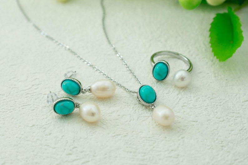 K&ouml;nnte beinhalten: Ein Schmuckset aus T&uuml;rkis und Perlen mit einer Halskette, Ohrringen und einem Ring. Die Halskette und die Ohrringe haben ovale T&uuml;rkis-Steine mit silbernen Fassungen, und der Ring hat einen T&uuml;rkis-Stein und eine Perle.