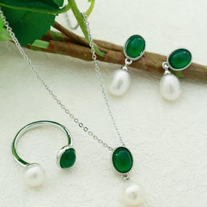 K&ouml;nnte beinhalten: Ein Schmuckset mit Halskette, Ring und Ohrringen mit einem gr&uuml;nen Edelstein und einem Perlenanh&auml;nger an jedem St&uuml;ck. Die gr&uuml;nen Steine sind oval geformt und in Silber gefasst.