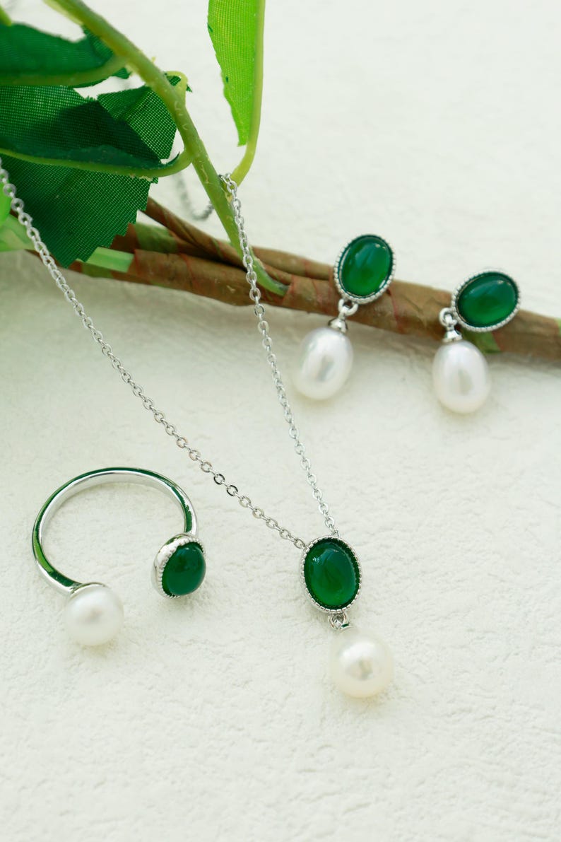 K&ouml;nnte beinhalten: Ein Schmuckset mit Halskette, Ring und Ohrringen mit einem gr&uuml;nen Edelstein und einem Perlenanh&auml;nger an jedem St&uuml;ck. Die gr&uuml;nen Steine sind oval geformt und in Silber gefasst.