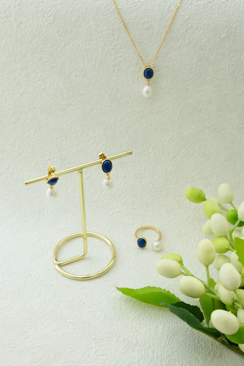 K&ouml;nnte beinhalten: Ein Satz Goldschmuck mit blauen und wei&szlig;en Steinen. Das Set beinhaltet eine Halskette, Ohrringe und einen Ring. Der Schmuck wird auf einem goldenen St&auml;nder vor einem wei&szlig;en Hintergrund pr&auml;sentiert.