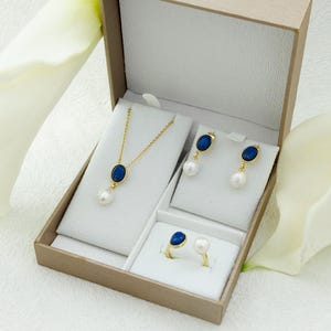 K&ouml;nnte beinhalten: Ein Schmuckset in einer wei&szlig;en Schachtel mit einem blauen ovalen Stein und einer Perle an jedem St&uuml;ck. Das Set enth&auml;lt eine Halskette, Ohrringe und einen Ring.