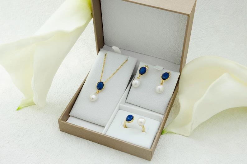 K&ouml;nnte beinhalten: Ein Schmuckset in einer wei&szlig;en Schachtel mit einem blauen ovalen Stein und einer Perle an jedem St&uuml;ck. Das Set enth&auml;lt eine Halskette, Ohrringe und einen Ring.