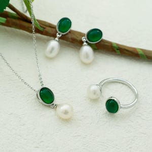 K&ouml;nnte beinhalten: Ein Halsketten-, Ohrring- und Ringset aus gr&uuml;nem Edelstein und Perle. Die Halskette und Ohrringe haben einen gr&uuml;nen ovalen Edelstein mit einer silbernen Fassung und eine wei&szlig;e Perle, die darunter h&auml;ngt. Der Ring hat einen gr&uuml;nen ovalen Edelstein auf der einen Seite und eine wei&szlig;e Perle auf der anderen.