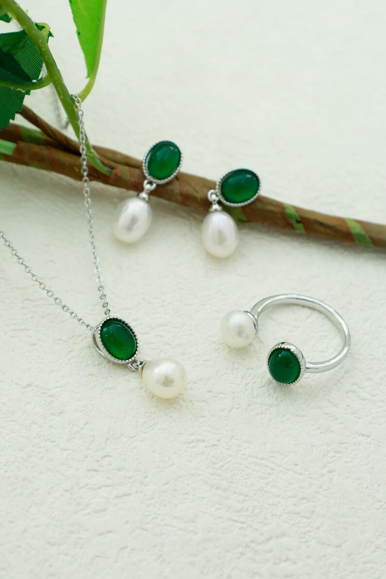 K&ouml;nnte beinhalten: Ein Halsketten-, Ohrring- und Ringset aus gr&uuml;nem Edelstein und Perle. Die Halskette und Ohrringe haben einen gr&uuml;nen ovalen Edelstein mit einer silbernen Fassung und eine wei&szlig;e Perle, die darunter h&auml;ngt. Der Ring hat einen gr&uuml;nen ovalen Edelstein auf der einen Seite und eine wei&szlig;e Perle auf der anderen.