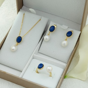 K&ouml;nnte beinhalten: Ein Gold-Set mit Halskette, Ohrringen und Ring mit blauen ovalen Steinen und wei&szlig;en Perlen. Der Schmuck ist in einer wei&szlig;en Schachtel ausgestellt.