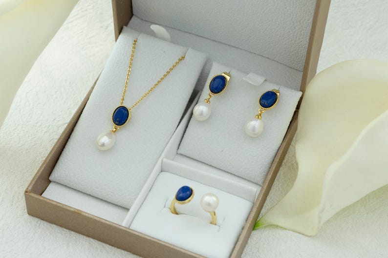 K&ouml;nnte beinhalten: Ein Gold-Set mit Halskette, Ohrringen und Ring mit blauen ovalen Steinen und wei&szlig;en Perlen. Der Schmuck ist in einer wei&szlig;en Schachtel ausgestellt.