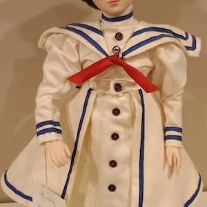Puede incluir: Muñeca de porcelana con un vestido de marinero color crema con ribetes azules y un lazo rojo. La muñeca lleva un sombrero de paja con una cinta roja. El vestido tiene botones marrones oscuros y una etiqueta con una ilustración.
