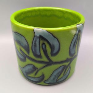 May include: A cylindrical ceramic planter with a glossy lime green glaze. The exterior features a hand-painted botanical motif consisting of a dark blue vine with stylised leaves wrapping around the circumference of the vessel.