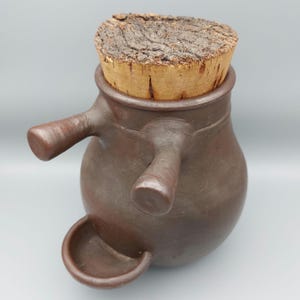 Peut inclure: Un pot en céramique marron avec un bouchon en liège. Le pot a deux poignées et un petit bec. Le bouchon en liège est de couleur marron clair et a une surface texturée. Le pot est de couleur marron foncé.