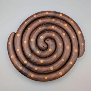 May include: A circular wooden trivet featuring a continuous spiral design crafted from dark brown wood. The surface is inlaid with small, evenly spaced light tan circular wooden dots that follow the curve of the spiral from the centre outwards.