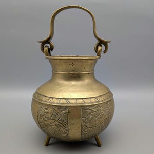 May include: A vintage brass pot with a handle and three small legs. The pot has an ornate design around the middle and a curved handle. The brass has a warm, aged patina, suggesting its age and use. The pot is likely used for decorative purposes.