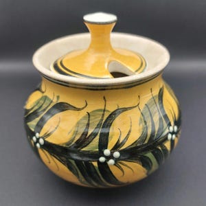 May include: A yellow ceramic sugar bowl with a lid. The bowl is decorated with black and green leaf-like patterns and small white dots. The lid has a handle and a slot for a spoon. The interior is a cream colour.