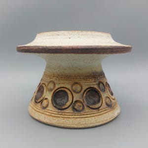 May include: A ceramic pedestal with a wide, flat top and a flared base. The top is off-white with a brown rim. The body is beige with a pattern of brown circles. The pedestal is likely used for displaying decorative items.