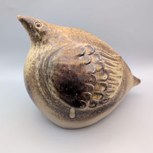 May include: A ceramic bird figurine with a rounded body and a speckled brown and beige glaze. The sculpture features a prominent wing with layered, scalloped feather details and a small, pointed beak, finished with a smooth, mottled texture.