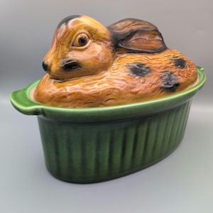 Op de afbeelding: Een keramische ovenschaal met deksel voorzien van een gebeeldhouwde bruine haas met donkere vlekken. De basis is een ovale, diepgroene schaal met verticale ribbels aan de buitenzijde en kleine handvatten aan beide uiteinden.