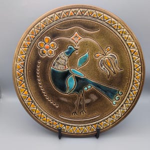 May include: A round ceramic plate with a brown glaze featuring a central stylised bird in shades of blue and teal. The design includes a flower, a geometric diamond shape, and a border of repeating orange and white triangles around the edge.