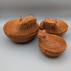 May include: Three brown woven wicker lidded containers shaped like frogs. Each container features a rounded body with a textured surface and two small, dark, glossy eyes positioned on the lid. The set includes one large and two smaller pieces.