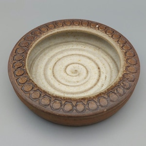 May include: A round ceramic bowl with a spiral pattern in the centre. The bowl has a light beige interior and a dark brown exterior with a repeating geometric pattern around the rim. The bowl is stoneware.