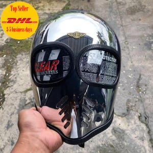 Può includere: Un casco da moto cromato con visiera nera e protezione per gli occhi. Il casco ha un'emblema dorato e il testo "CLEAR SEE YOUR EYES" sulla visiera. Il casco è tenuto da una mano.
