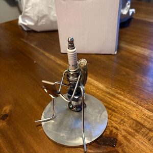 以下が含まれることがあります： スパークプラグと金属線で作られた、椅子に座っている人のユニークな金属彫刻。その人物は円形の金属ベースに座っています。この彫刻は創造的な芸術作品です。