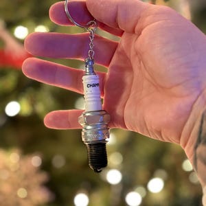 Op de afbeelding: Een sleutelhanger met een Champion bougie. De bougie is wit met het woord "CHAMP" erop gedrukt. Hij heeft een zilverkleurige metalen basis en een zwarte punt. De sleutelhanger is bevestigd aan een zilveren metalen ring.