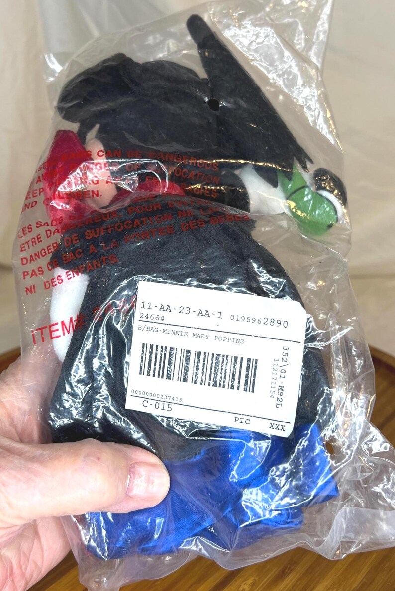 K&ouml;nnte beinhalten: Eine Minnie Mary Poppins Puppe in einer Plastikt&uuml;te. Die Puppe hat einen schwarzen Hut, schwarze Haare, eine rote Schleife und ein blaues Kleid. Die Verpackung enth&auml;lt ein Produktetikett mit Barcode und Artikelnummer.