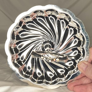May include: A silver-colored, decorative serving dish with a scalloped edge and a swirling, fan-like pattern. The dish has a small handle with iridescent accents. The dish is round and appears to be made of metal.