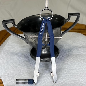 Puede incluir: Un compás azul y plateado con el nombre de la marca "STAEDTLER" impreso en el lateral. El compás está sobre una superficie blanca junto a una copa de plata.