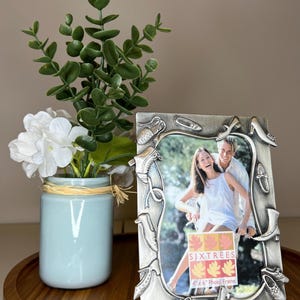 May include: A decorative silver photo frame with shoe embellishments surrounds a photo of a couple. A small blue vase with white flowers and greenery sits to the left. The frame is designed for a 4x6 inch photo.