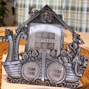 May include: A silver-toned metal picture frame shaped like a boat with animal figures. The frame has two openings for pictures and the words "Sixtrees" are printed on the front.
