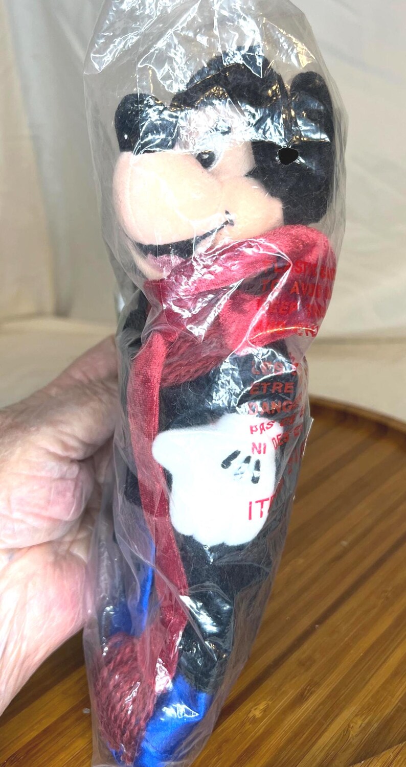 K&ouml;nnte beinhalten: Ein Mickey Mouse Pl&uuml;schtier in einer durchsichtigen Plastikt&uuml;te. Das Pl&uuml;schtier hat einen schwarzen K&ouml;rper, einen roten Schal und wei&szlig;e Handschuhe. Die Verpackung hat Text.