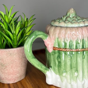 Op de afbeelding: Een decoratieve theepot en een kleine potplant. De theepot is groen, roze en wit, met een handvat en een tuit. De theepot is ontworpen om op asperges te lijken. De plant is groen en in een roze pot.
