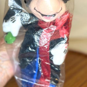 K&ouml;nnte beinhalten: Ein Mickey Mouse Pl&uuml;schtier in einer durchsichtigen Plastikt&uuml;te. Das Pl&uuml;schtier hat einen schwarzen Kopf und Ohren, einen roten Schal und eine blaue Hose. Es h&auml;lt einen gr&uuml;nen Gegenstand in seiner wei&szlig;en behandschuhten Hand.