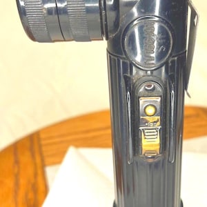 Puede incluir: Una linterna negra en ángulo recto con un mango acanalado y una cabeza texturizada. La linterna tiene un interruptor amarillo y un clip metálico. El texto "U.S. MX-991/U" es visible en el lateral.