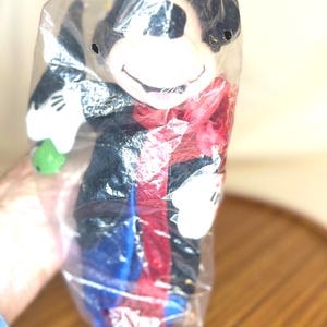K&ouml;nnte beinhalten: Ein Mickey Mouse Pl&uuml;schtier in einer durchsichtigen Plastikt&uuml;te. Die Puppe hat einen schwarzen K&ouml;rper, wei&szlig;e Handschuhe und einen roten Schal. Es h&auml;lt einen gr&uuml;nen Gegenstand. Das Gesicht der Puppe ist wei&szlig; mit einem l&auml;chelnden Ausdruck.