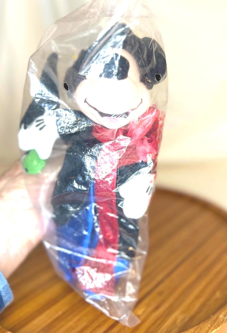 K&ouml;nnte beinhalten: Ein Mickey Mouse Pl&uuml;schtier in einer durchsichtigen Plastikt&uuml;te. Die Puppe hat einen schwarzen K&ouml;rper, wei&szlig;e Handschuhe und einen roten Schal. Es h&auml;lt einen gr&uuml;nen Gegenstand. Das Gesicht der Puppe ist wei&szlig; mit einem l&auml;chelnden Ausdruck.