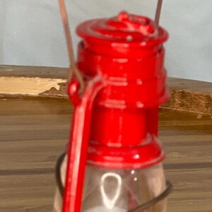 May include: A red metal oil lamp with a clear glass globe and a wire handle. The lamp is sitting on a wooden surface.