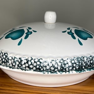 以下が含まれることがあります： 白のセラミック製サービングディッシュ。蓋にはティール色の花柄、縁には水玉模様のスカラップ模様が施されています。蓋には小さな白いノブが付いています。木製の表面に置かれています。