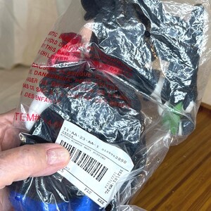 K&ouml;nnte beinhalten: Eine Minnie Mouse Pl&uuml;schpuppe in einer Plastikt&uuml;te. Die Puppe tr&auml;gt einen schwarzen Hut, ein schwarzes Oberteil und einen blauen Rock. Ein Etikett mit Barcode und Text ist an der T&uuml;te befestigt. Die Puppe ist teilweise sichtbar.